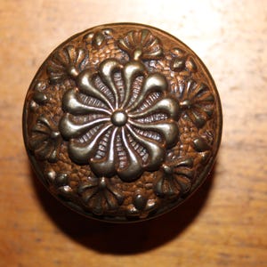 May include: Ornate, antique bronze cabinet knob with a floral design. The knob features a central flower surrounded by smaller floral accents, all set against a textured background. A vintage hardware piece.