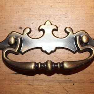 May include: A decorative brass drawer pull with a ornate design. The pull has a silver finish and a curved shape.