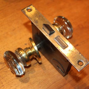 May include: Antique door hardware set featuring a brass strike plate and two clear glass doorknobs. The set includes a black metal lock box and brass hardware. The doorknobs have a ribbed design.