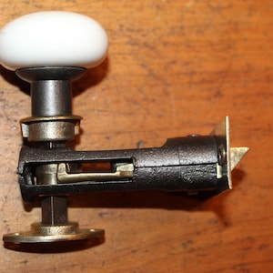 May include: A vintage door knob with a white porcelain handle and a black metal base. The knob is attached to a brass mechanism with a keyhole.