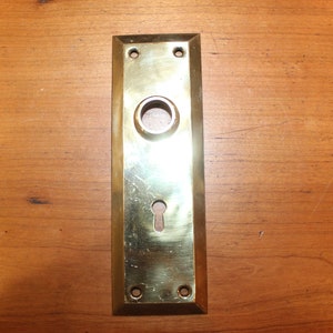 May include: A brass door plate with a keyhole and a round hole for a doorknob.