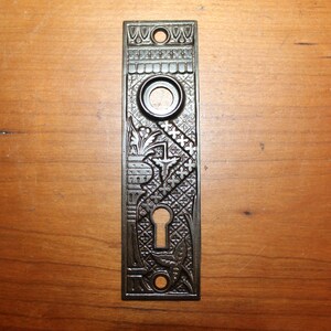 May include: A vintage, ornate, cast iron door plate with a keyhole and a decorative design. The plate is a dark, aged silver color.