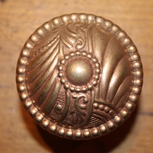 May include: Ornate, antique-style brass knob with a detailed, raised floral and scroll design. The knob features a central circular element surrounded by a beaded border and is likely for a cabinet or drawer.