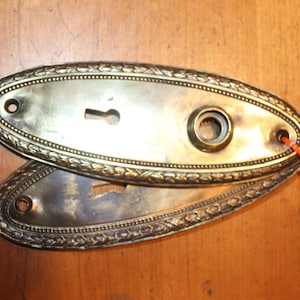 May include: Two antique brass door plates with keyholes and decorative edging. The plates are oval-shaped and show signs of age and wear. An orange string is attached to one plate. The plates are likely for a vintage door.