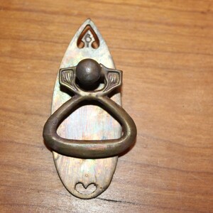 May include: A vintage brass door pull with an oval-shaped backplate and a decorative ring handle. The handle has a round knob at the top and intricate floral designs.