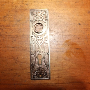 May include: Antique bronze-toned door plate with ornate floral and geometric designs. Features a keyhole, a circular opening, and decorative embellishments. The rectangular plate is approximately 8 inches long.
