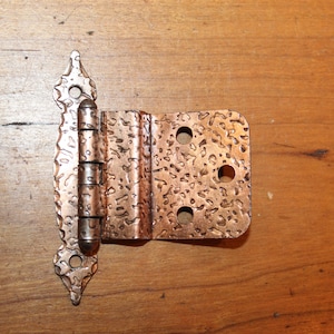 May include: A copper-colored hinge with a textured surface and decorative edges. The hinge is open, revealing the barrel and pin. The hinge is made of metal and has three holes on the rectangular side. The hinge is on a wooden surface.