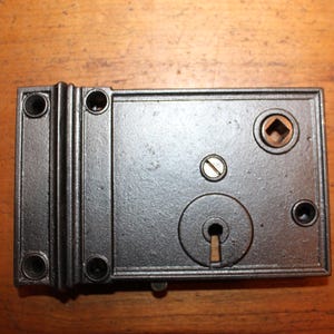 May include: A black metal door lock with a keyhole and a small square hole for a screwdriver. The lock has a rectangular shape and is made of cast iron.