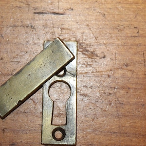 May include: A brass keyhole cover with a rectangular plate and a separate flap that covers the keyhole. The flap is hinged and can be lifted to reveal the keyhole.