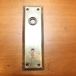May include: A brass door plate with a keyhole and a round hole for a doorknob. The plate has a worn, aged look.