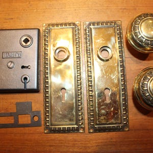 May include: Antique door hardware set including a dark metal mortise lock, two brass door plates, and two brass doorknobs. The doorknobs have a decorative circular pattern. The set is displayed on a wooden surface.