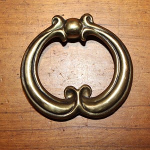 May include: A vintage brass drawer pull with an ornate, oval design. The pull features a central sphere and decorative scrollwork at the top and bottom. The hardware is set against a wooden surface.