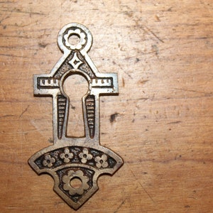 May include: A decorative keyhole cover made of metal with an ornate design. The cover is shaped like a cross with a keyhole in the center. The keyhole is surrounded by a decorative border. The cover is finished in a brass color.