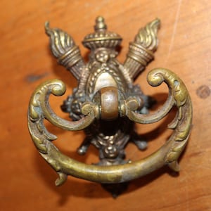 May include: A vintage brass cabinet pull with a decorative design featuring a central crest with two stylized torches. The pull has a curved handle and ornate details.