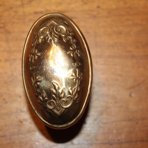 May include: A vintage, oval-shaped brass door knob with an ornate floral design. The knob is a warm gold color and features a detailed embossed pattern of leaves and flowers. The design is centered on the knob's surface.