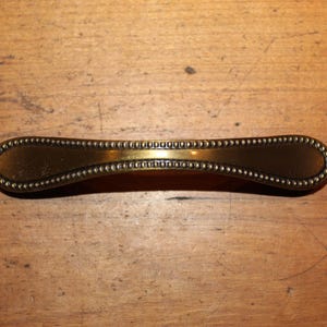 May include: A gold-toned cabinet pull with a beaded edge and a slightly curved shape.