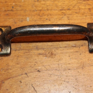 May include: A vintage-style metal handle with a curved grip and two mounting plates. The handle has a dark, aged finish with visible wear, and each plate features two circular holes for screws. The handle is likely for a cabinet or drawer.