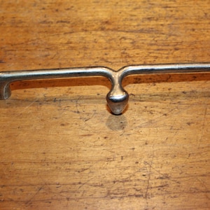 May include: A silver-toned metal cabinet pull handle with a distinctive design. The handle features a central, rounded detail and curved ends for easy gripping. The hardware is set against a warm, wooden surface.