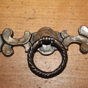 May include: A decorative brass drawer pull with a ring handle. The pull has a scrolled design and a dark brown finish.