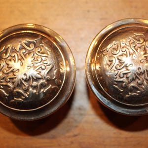 May include: Two antique brass door knobs with ornate floral designs. The knobs are round with a slightly domed top and a detailed pattern of leaves and swirls. The brass has a warm, aged patina.