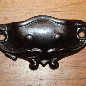 May include: Black metal drawer pull with a decorative design. The pull has two holes for mounting.