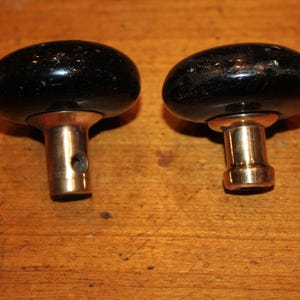 May include: Two vintage black oval door knobs with brass stems. The knobs are set against a warm, wooden surface. The knobs have a glossy finish and show signs of wear, suggesting age and use. Hardware for doors.