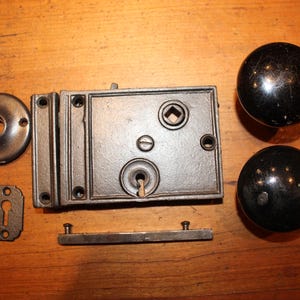 May include: An antique door lock set with a rectangular lock body, a round door plate, a keyhole cover, a strike plate, a bar, and two black spherical door knobs. The set is made of metal and is displayed on a wooden surface.