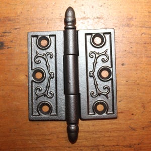 May include: Black cast iron hinge with a decorative scroll design. The hinge has a pointed top and bottom.