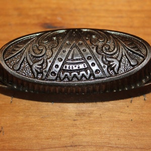 May include: A black metal drawer pull with an ornate design. The pull has a rounded oval shape and is decorated with a geometric pattern and floral motifs.