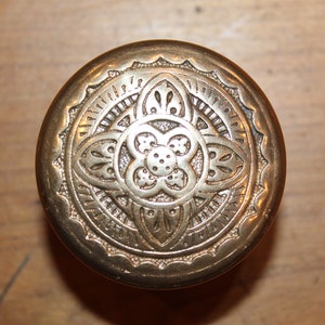 May include: A round, antique brass door knob with an ornate floral design. The knob features a central flower motif surrounded by detailed patterns and a decorative edge. The brass has a warm, aged patina.