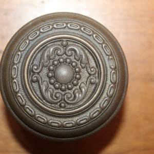 May include: A vintage-style door knob with a decorative design featuring a central circle surrounded by intricate floral patterns. The knob is made of brass and has a dark, aged finish.