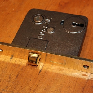 May include: A vintage, rectangular mortise lock with a brass faceplate and a dark metal body. The lock features a keyhole, a bolt, and screw holes for mounting. The brass faceplate extends horizontally, with a rectangular bolt and two screw holes.