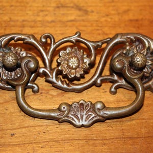 May include: Ornate antique bronze drawer pull with floral and scrollwork details. The handle features a central flower and two flower-shaped accents on either side. A decorative piece for furniture restoration.
