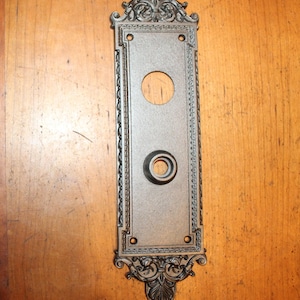 May include: An antique bronze-colored door plate with ornate scrollwork and a rectangular shape. The plate features a large circular opening at the top and a smaller one at the bottom, likely for a doorbell or button. The edges are decorated with a detailed rope-like pattern.