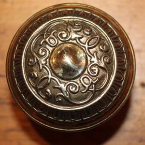May include: Close-up of a vintage brass doorknob. The knob features an ornate, embossed design with a central circular element and swirling patterns. The brass has a warm, aged patina, suggesting its antique nature.