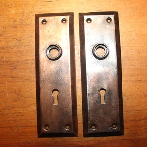 May include: Two antique rectangular door plates with a keyhole and a circular opening for the doorknob. The plates are made of a dark metal with a distressed finish, showing signs of age and wear. Each plate has four screw holes.