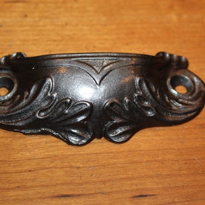 May include: Black metal drawer pull with a decorative, ornate design. The pull has a curved shape and a smooth, slightly shiny finish.