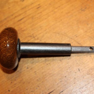 May include: A wooden cabinet knob with a dark brown, glossy finish and a metal shaft. The knob has a rounded shape with visible wood grain. The metal shaft is cylindrical with a square end and a small hole.