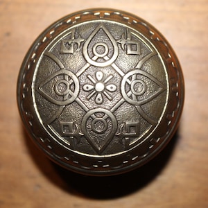 May include: A round, brass door knob with an intricate, embossed design. The design features a central flower with four surrounding eye-like shapes.