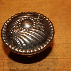 May include: A silver-toned cabinet knob with an oval shape. The knob features a central circular design surrounded by a beaded border and ornate leaf-like patterns. The hardware is likely made of metal.