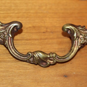 May include: A brass drawer pull with a decorative, ornate design. The pull is shaped like a U with intricate floral details.
