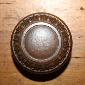 May include: A vintage bronze door knob with a circular design. The knob features a central, slightly raised disc surrounded by a decorative border of repeating patterns. The metal has a weathered appearance, suggesting age and use.