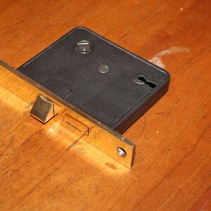 May include: A vintage door lock with a black rectangular body and a gold-colored metal plate. The lock features a keyhole and is mounted on a wooden surface. The metal plate has a rectangular shape with two screw holes.