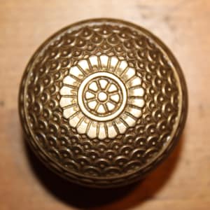 May include: Ornate, antique brass doorknob with a detailed, circular floral design at the center. The knob has a textured surface with a repeating pattern of small, curved shapes. The overall color is a warm, aged brass.