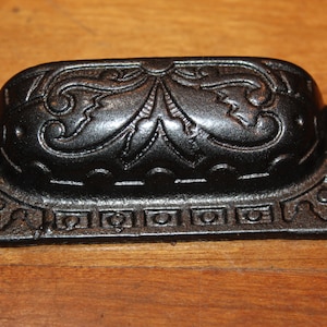 May include: Black cast iron drawer pull with an ornate floral design. The pull has a rectangular base with rounded edges and a raised oval center.