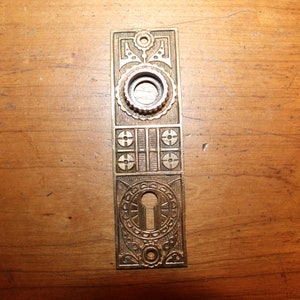 May include: A brass door plate with an ornate design. The plate has a keyhole and a circular knob. The plate is mounted on a wooden surface.
