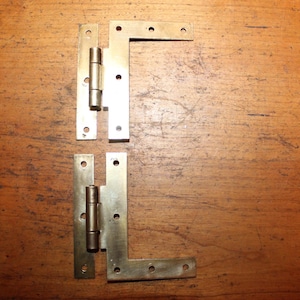 May include: Two brass hinges with a 90-degree angle. The hinges are attached to a wooden surface.