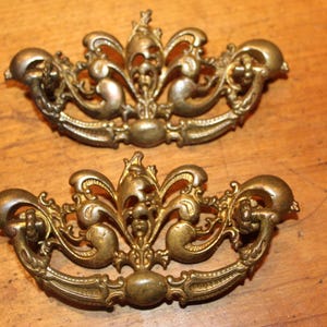 May include: Two antique brass drawer pulls with ornate, floral designs. The pulls have a curved handle and decorative embellishments, perfect for adding a vintage touch to furniture. The brass has a warm, aged patina.