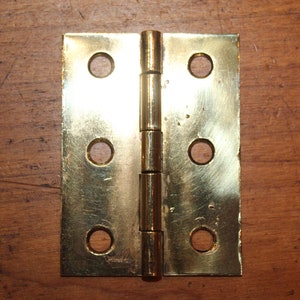 May include: A pair of brass hinges with four holes on each side. The hinges are closed and ready to be installed.