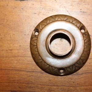 May include: A vintage brass door knob backplate with a central opening and decorative leaf pattern around the edge. The backplate has a circular shape and three screw holes. The finish is aged and tarnished.